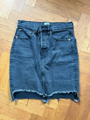 Svart jeanskjol - Cool svart jeanskjol med rå, fransig nederkant och klassiska fickor fram och bak. Kjolen har knappgylf och bälteshällor, perfekt för en avslappnad streetstyle-look.