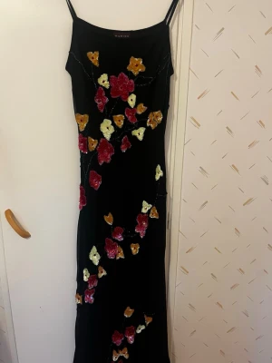 Svart långklänning med blommor - Svart långklänning med tunna axelband och blommor i rött, gult och vitt. Klänningen har en smal passform och blommorna är dekorerade med paljetter för extra effekt. Perfekt för dig som vill sticka ut med en unik och elegant look. strechigt material så kan passa S/M/L. Använd ett fåtal gånger sparsamt och köpt utomlands. 