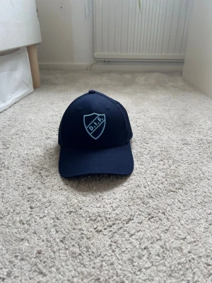 Mörkblå DIF keps med broderad logga - Snygg mörkblå keps med broderad DIF-logga i ljusblått framtill. Klassisk böjd skärm och justerbar baksida. Perfekt för dig som vill visa ditt stöd för Djurgården. Tillverkad i ett slitstarkt tygmaterial.