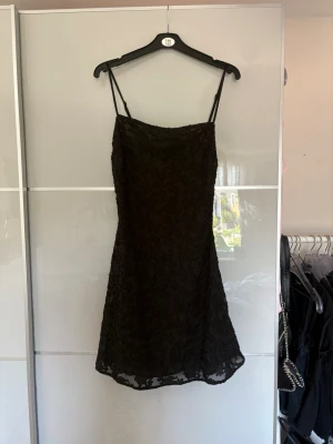 Svart spetsklänning med smala axelband - Snygg svart kort klänning med smala axelband och spetsdetaljer. Klänningen har en lätt utsvängd passform och är perfekt för dig som gillar en stilren look med lite extra textur. Fodrad under spetsen för en bekväm känsla.