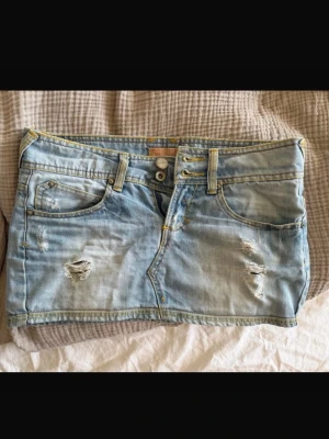 Ljusblå jeanskjol med slitningar - Säljer en ljusblå jeanskjol i kort modell med slitna detaljer framtill och bakfickor. Kjolen har klassisk femficksdesign, bälteshällor och dragkedja med knapp framtill. Perfekt för en avslappnad och trendig look.