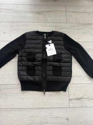 Svart cardiganjacka från Moncler - Snygg svart cardiganjacka från Moncler med quiltad front och två ribbade fickor. Jackan har rund halsringning, dragkedja framtill och mjuka ärmar i sweatshirtmaterial. Perfekt för lager på lager och streetstyle.