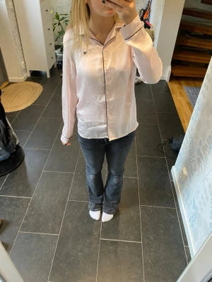 Ljusrosa pyjamasskjorta med svarta kanter - Säljer en ljusrosa pyjamasskjorta med svarta kontrastsömmar och knappar framtill. Skjortan har lång ärm och klassisk krage, tillverkad i ett glansigt, mjukt material som känns lyxigt mot huden. Perfekt för mysiga kvällar hemma.