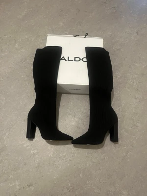Svarta knähöga stövlar från ALDO - Snygga svarta knähöga stövlar från ALDO med ribbad textur och bred klack. Stövlarna har spetsig tå och är tillverkade i ett stretchigt tygmaterial som sitter tight runt benet. Perfekta för att lyfta vilken outfit som helst. Helt nya☺️☺️