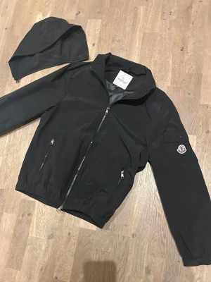 Svart vindjacka - ”KAN DISKUTERA PRIS” Snygg svart vindjacka från Moncler med avtagbar huva och dragkedja framtill. Jackan har två fickor med dragkedja och en Moncler-logga på ärmen. Tillverkad i ett lätt och vattenavvisande material, perfekt för blåsiga dagar.