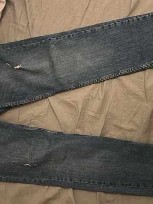 Mörkblå raka jeans med slitningar - Säljer ett par mörkblå jeans med raka ben och slitna detaljer vid knäna. Jeansen har en klassisk femficksmodell och är tillverkade i denim. Perfekta för en avslappnad och trendig look.