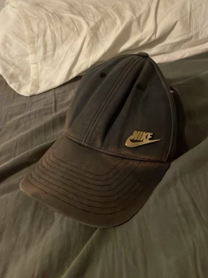 Svart Nike keps med guldfärgad logga - Snygg svart keps från Nike med böjd skärm och guldfärgad Nike-logga framtill. Klassisk sexpanel-design och justerbar passform. Tillverkad i bomullsmix för en bekväm känsla. Perfekt accessoar för en sportig och avslappnad stil.