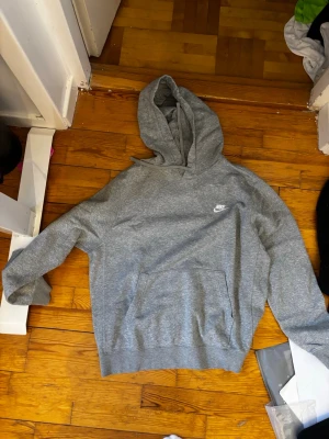 Grå hoodie från Nike med logga - Klassisk grå hoodie från Nike med vit broderad logga på bröstet. Hoodien har lång ärm, stor huva och magficka framtill. Tillverkad i mjuk bomullsmix och passar perfekt för en avslappnad stil.