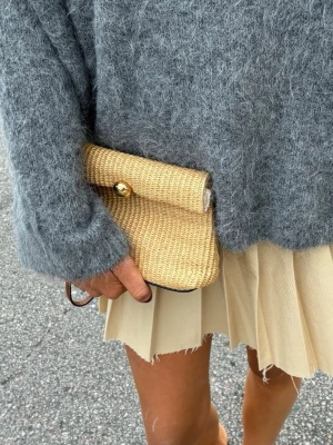 Beige flätad kuvertväska i strå - Snygg beige kuvertväska i flätat stråmaterial med guldfärgad knappstängning. Väskan har en enkel och stilren design som passar perfekt till olika outfits. Liten och smidig att bära i handen.