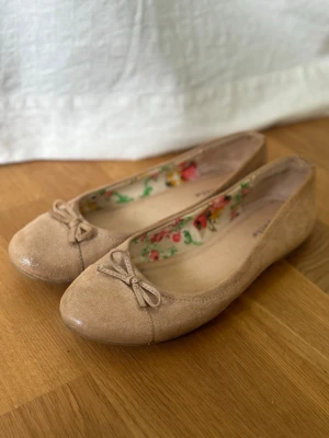 Beige ballerinaskor från Depeche - Snygga beige och glittriga ballerinaskor från Depeche i mockaimitation. Skorna har en liten rosett framtill och insidan är fodrad med ett blommigt tyg i färgglada nyanser. Perfekta för dig som gillar en söt och stilren look.