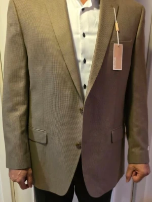 Beige kavaj med diskret mönster - Stilren beige kavaj med diskret småmönstrad struktur. Klassisk passform med två knappar framtill, två fickor med lock och en bröstficka. Fodrad insida och innerficka. Perfekt till skjorta och svarta byxor för en snygg look.