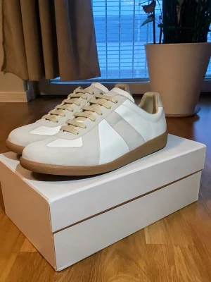 Maison Margiela Gats - Maison Margiela sneakers i vitt och beige med klassisk gummisula. Ovandelen är i en mix av slätt läder och mocka, med beige detaljer och vita paneler. Låga i modellen och med beige snörning. Perfekta för en clean och stilren look.