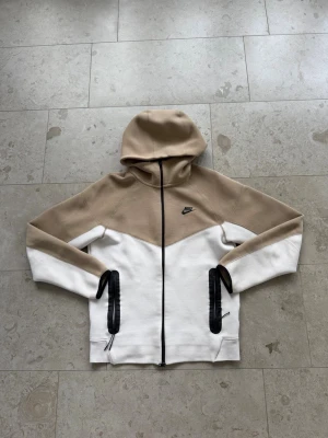 Nike beige och vit hoodie med dragkedja - Nike hoodie i beige och vitt med svart logga på bröstet. Hoodien har hel dragkedja framtill, två svarta dragkedjeförsedda fickor och en stor huva. Tillverkad i mjukt bomullsmaterial, perfekt för en avslappnad streetwear-look.