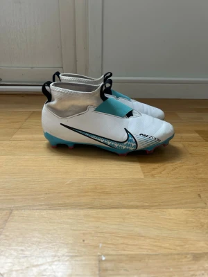 Nike Air Zoom vita fotbollsskor  - Säljer ett par vita Nike Air Zoom fotbollsskor med turkosa och svarta detaljer samt rosa dobbar. Skorna har en strumpliknande passform utan snörning och coolt mönster på sidan. Perfekta för dig som vill sticka ut på planen. PRISET KAN DISKUTERAS!