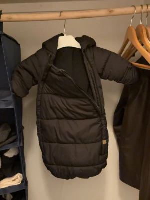 Svart dunoverall med huva - Säljer en svart dunoverall med huva och långa ärmar. Overallen har en sned dragkedja framtill och är vadderad för extra värme. Perfekt för kalla vinterdagar och håller dig riktigt varm.