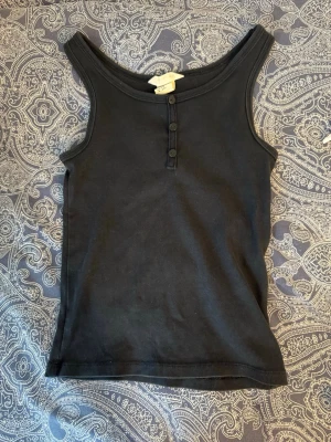 Svart ribbad linnetopp med knappar - Säljer en svart ribbad linnetopp med rundad halsringning och tre knappar framtill. Toppen är ärmlös och har en enkel, stilren design som passar till det mesta. Perfekt att matcha med jeans eller kjol för en avslappnad look.