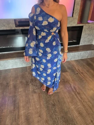 Blå blommig oneshoulder klänning - Snygg blå långklänning med vita och gula blommor. Klänningen har oneshoulder-design med en lång, vid ärm och en axel bar. Volangdetaljer längs sidan och en dekorativ ring i midjan. Tillverkad i ett lätt, flowy material som känns som polyester.