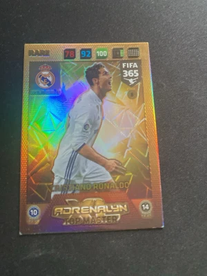 Panini Adrenalyn FIFA 365 Top Master kort - Säljer ett glittrigt Panini Adrenalyn XL FIFA 365 Top Master samlarkort med Cristiano Ronaldo i Real Madrid-tröja. Kortet har färgglad holografisk yta och visar stats för Defence, Control och Attack. Perfekt för samlare av fotbollskort.
