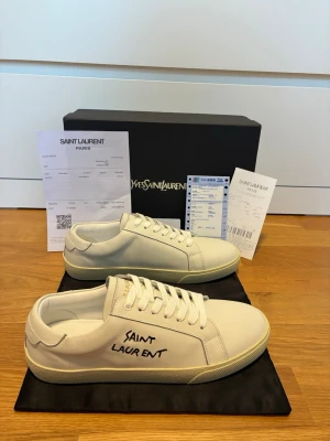 Saint Laurent sneakers - Säljer ett par vita sneakers från Saint Laurent med handskriven logga på sidan. Skorna har klassisk låg profil, vita snören och sula i gummi. Tillverkade i slätt läder och levereras med originalkartong och dustbag.