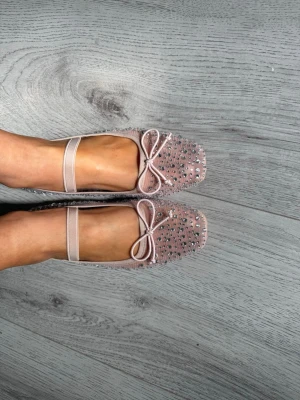 Nude ballerinaskor med strass och rosett - Säljer ett par ljusrosa/Nude  ballerinaskor med elastiskt band över foten, dekorerade med små glittriga strass och en söt rosett framtill. Skorna har rund tå och är tillverkade i ett mesh-liknande material som ger en luftig känsla.