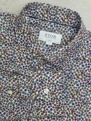 Eton Slim skjorta blommig flerfärgad storlek L - Blommig skjorta från Eton i Slim-passform. Småblommigt flerfärgat mönster i bland annat blått, grönt, rött, orange och rosa på vit botten. Tillverkad i 100% bomull i poplinkvalitet.  Storlek L / 41 / 16 Slim Fit  Mycket fint skick.  Mått • Bröstbredd: ca 54 cm • Plagglängd: ca 78 cm • Axelbredd: ca 49 cm • Ärmlängd: ca 68 cm  Material 100% bomull  Varan är begagnad och säljs i befintligt skick, se bilder för detaljer.  Tycker du priset är för högt? Skicka ett prisförslag!