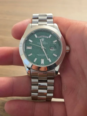 Stilren klocka med grön urtavla - Stilren klocka med grön urtavla och dag samt datumvisning. Klockan har en klassisk rund form och ett robust armband i silverfärgat metall. Indexmarkeringarna är rektangulära och boetten har en blank finish som ger en exklusiv känsla.
