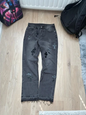 Grå jeans med svarta korspatchar - Säljer ett par grå jeans med raka ben och råa kanter nertill. Jeansen har flera svarta korspatchar i läderlook på framsidan som ger en edgy vibe. Materialet är klassisk denim och passformen är relaxed. Perfekta för dig som gillar streetwear och unika detaljer.
