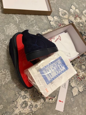 Mörkblå sneakers med röd sula - Snygga mörkblå sneakers från Christian Louboutin i mocka med klassisk röd sula. Skorna har en stilren design med diskreta sömmar och svarta snören. Perfekta för dig som vill sticka ut med exklusiva detaljer och lyxig känsla.