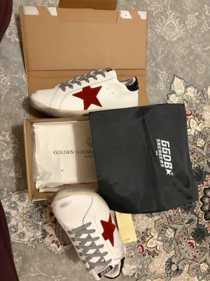Golden Goose vita sneakers med röd stjärna - Golden Goose sneakers i vitt läder med en ikonisk röd stjärna på sidan och svarta detaljer vid hälen. Skorna har grå skosnören och en vintage-inspirerad sula. Perfekta för dig som gillar en clean men ändå unik look.