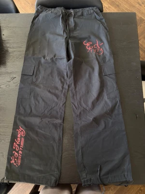Svarta cargobyxor från Ed Hardy - Snygga svarta cargobyxor från Ed Hardy med röda tryckdetaljer och logga på benet och höften. Byxorna har flera fickor på sidorna och dragsko i midjan samt vid bensluten. Perfekta för en streetwear-look.