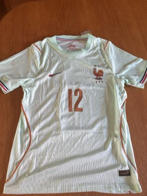 Frankrike fotbollströja Barcola 12 - Ljus fotbollströja för Frankrikes landslag med nummer 12 och namnet Barcola på ryggen. Tröjan har korta ärmar, tunna ränder, broderad tupp-logga och Nike-märke. Detaljer i rött och blått på ärmsluten samt orangea linjer längs sidorna. Tillverkad i lätt funktionsmaterial.