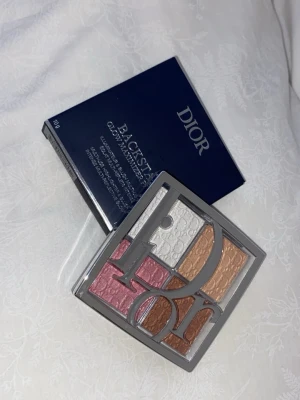 Dior Backstage Glow Face Palette - Dior Backstage Glow Face Palette med fyra skimrande nyanser: rosa, vit, guld och koppar. Paletten har en stilren silverfärgad förpackning med genomskinligt lock och stor Dior-logga. Perfekt för att skapa glow och highlighta ansiktet.
