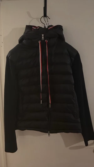 Svart pufferjacka från Moncler - Snygg svart pufferjacka från Moncler med huva och dragkedja framtill. Jackan har röda och vita detaljer på snörena och Moncler-logga på ärmen. Tillverkad i vadderat material som håller värmen under kalla dagar. Perfekt för dig som vill ha både stil och funktion.