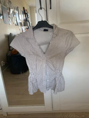Randig kortärmad skjorta med knappar - Snygg vit skjorta med smala svarta ränder och korta ärmar. Skjortan har knappar framtill, krage och insydd resår i midjan för en mer markerad siluett. Perfekt till jeans eller kjol för en fräsch look.