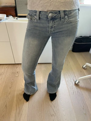 Ljusblå bootcut jeans True Religion - Säljer ett par ljusblå bootcut jeans från True Religion med klassiska sömmar och detaljerade bakfickor med lock och knappar. Jeansen har normal midja och är tillverkade i denim med lätt stretch för skön passform. Perfekta till en trendig stil. Säljer då jag inte tycker om hur de sitter på mig. Modellen är 166 cm lång. (Byxorna är uppsydda)☺️