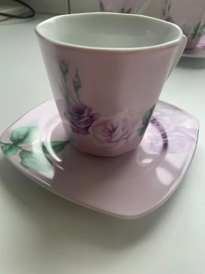 Rosa kaffekoppar med fat och blommor - Detta är en uppsättning kaffekoppar med fat, sannolikt från serien Mariehult Sweden, kända för sin fyrkantiga form och romantiska rosmotiv.  Material: Porslin. Design: Ljusrosa med lila rosor och gröna blad. Form: Mjukt fyrkantiga fat och matchande koppar. Användning: Passar för kaffe eller te.   Är ej använda, legat i skåp.   Priset är för hela uppsättning inkl 7 koppar och 7 fat.