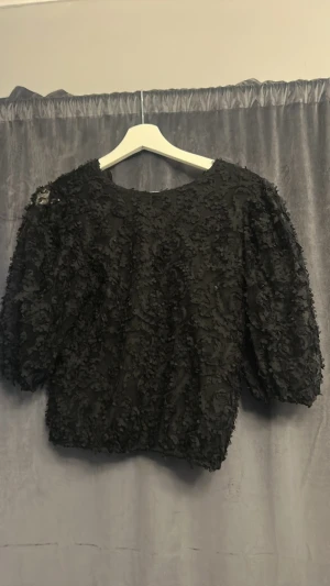 Svart blus med blomspets - Säljer en svart blus med puffiga ärmar och 3D-volangdetaljer över hela tyget. Blusen har rund halsringning och en croppad passform som ger en trendig look. Perfekt för dig som vill sticka ut lite extra.