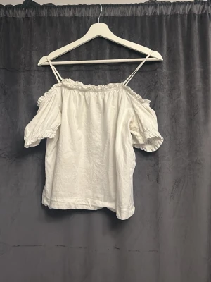 Singoalla blus - Söt vit offshoulder blus med tunna axelband och volangdetaljer vid ärmslut och halsringning. Blusen har korta puffärmar och en luftig passform, perfekt för varma dagar. Tillverkad i mjukt bomullstyg.