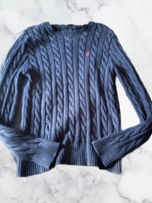 Ralph lauren kabelstickad tröja - Kabelstickad tröja i mörkblått med rund hals och långa ärmar. Tröjan har ett diskret rött broderat märke på bröstet och ribbade muddar vid ärmslut och nederkant. Perfekt för dig som gillar klassisk och stilren look.