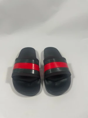 Guccislides - Snygga svarta sandaler i slides-modell med breda remmar och en tydlig röd rand över foten. Tillverkade i syntetmaterial med räfflad sula för extra grepp. Perfekta för en avslappnad och trendig look.