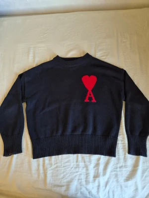 Ami paris Svart - Svart stickad tröja med rund hals och ribbade muddar. På bröstet finns ett stort rött hjärta med bokstaven A under, vilket ger en unik och trendig look. Tröjan har en avslappnad passform och är perfekt för dig som gillar stilrena detaljer.