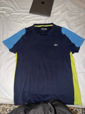 Blå Lacoste t-shirt med stor logga - Säljer en snygg t-shirt från Lacoste i mörkblått med ljusblå ärmar och limegröna sidopaneler. Framtill finns den klassiska lilla krokodilloggan och på ryggen en stor vit krokodil. Perfekt för dig som gillar sportig stil.