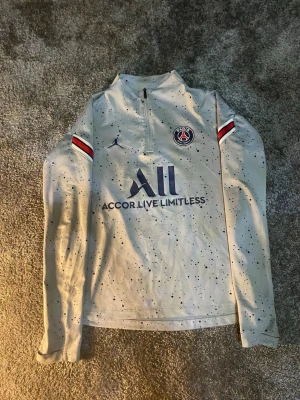Grå PSG träningströja med tryck - Snygg grå långärmad träningströja från PSG med svartprickigt mönster, halv dragkedja och klubbmärke på bröstet. Tröjan har tryckta detaljer som 'Accor Live Limitless' och 'Visit Rwanda' samt rödvita ränder på ärmarna. Perfekt för fotbollsträning eller chill.