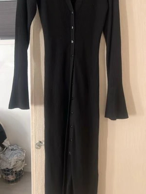 Svart ribbad långklänning - Snygg svart långklänning med ribbad struktur och långa ärmar. Klänningen har knappar hela vägen framtill och en smickrande v-ringning. Perfekt för dig som gillar en stilren och enkel look.