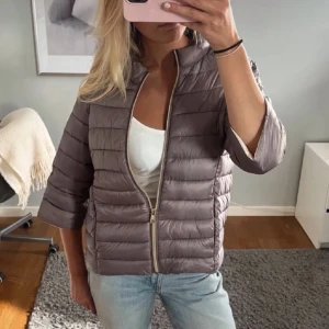 Ljusbrun pufferjacka med dragkedja - Säljer en ljusbrun pufferjacka med horisontella sömmar och dragkedja framtill. Jackan har en hög krage och trekvartsärmar, vilket ger en avslappnad och trendig look. Perfekt för kalla dagar när du vill vara både varm och snygg.💓