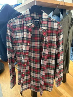 Rutig skjorta från GAP - Klassisk rutig skjorta från GAP i bomull. Skjortan har en vit bas med röda, gröna och blå rutor, knappar framtill och lång ärm. Perfekt för dig som gillar en tidlös och avslappnad stil.
