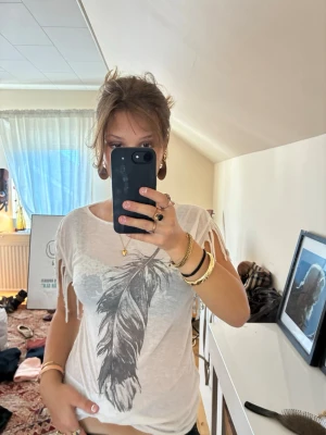 Vit t-shirt med fjädertryck och fransar - Säljer en vit t-shirt med stort fjädertryck framtill och fransdetaljer längs ärmarna. T-shirten har rund halsringning och är kortärmad. Materialet känns lätt och luftigt, perfekt för varmare dagar.