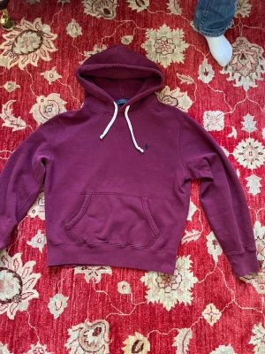 Vinröd hoodie med ficka och huva - Säljer en vinröd hoodie med klassisk känguruficka och vit dragsko i huvan. Hoodien har ett litet broderat märke på bröstet och ribbade muddar vid ärmar och nederkant. Perfekt för chill dagar eller när du vill vara extra bekväm. Ny pris ca 2700