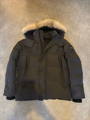 Svart dunjacka med huva och fuskpäls - Snygg svart dunjacka med huva och fuskpälsdetalj. Jackan har en quiltad design, dragkedja och knappar framtill samt två stora fickor. Muddar vid ärmslut för extra värme och en patch på ärmen för en cool touch.