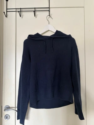 Mörkblå stickad hoodie - Mysig mörkblå stickad hoodie med huva och dragsko. Tröjan har långa ärmar och bred ribbad kant nedtill samt vid ärmsluten. Perfekt för dig som gillar en enkel och stilren look med en avslappnad passform.